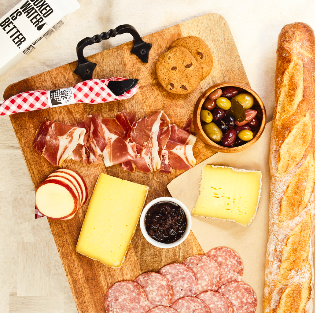 Cured meats, cheese, baguette, olives, cookies, and jam on a wooden cutting board