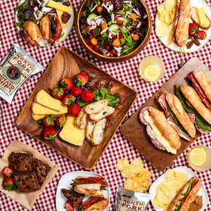 Assorted picnic foods including baguettini sandwiches, salads, cheese, and desserts