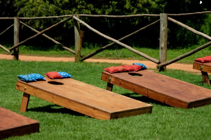 Cornhole boards with red and blue bean bags on grass near wooden fence.